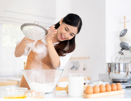 Young woman wearing light brown apron in the modern kitchen have fun sifting bread flour, preparing...