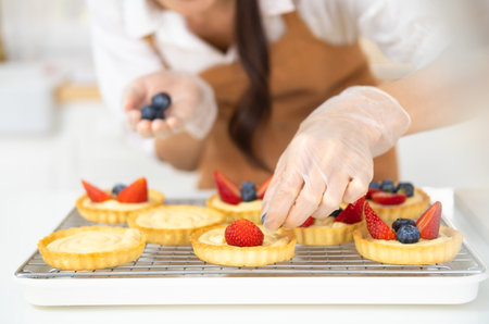 Young beautiful woman is baking in kitchen , bakery and coffee shop business. Hands in cooking gloves decorating freshly baked tart with blueberries strawberry fresh fruit. homemade bakery at home.の写真素材