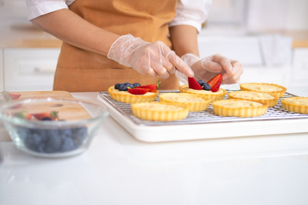close up of hands in cooking gloves Baker adding blueberries strawberry fresh fruit to a tart on white table in Kitchen. housewife baker wear apron making fruit tart. homemade bakery at home.の写真素材