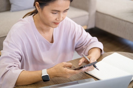 Young business woman sitting at table and taking notes in notebook,holding smartphone. On table is...
