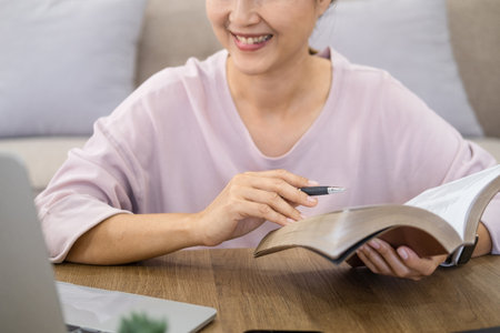 Focused young asian businesswoman or student looking at laptop holding book learning, serious woman working or studying with computer doing research or preparing for exam online.の写真素材