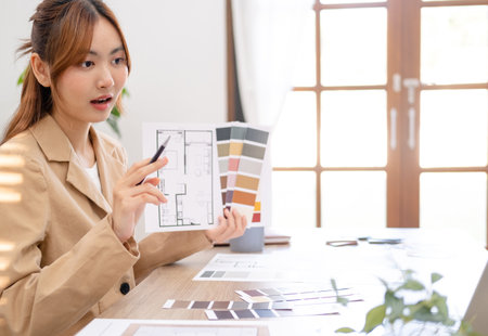 Interior designer talking with client on video call, showing architecture blueprint of residence. Asian architect woman sitting in office, looking at laptop display with online conference.の写真素材