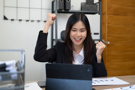 Sale of online asian woman special she is satisfied with sales success. Successful business woman with arms up celebrating. Excited asian woman sit at desk feel euphoric win online lottery.の写真素材