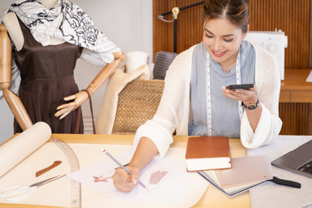 Young attractive asian female fashion designer leaning on office desk, talking on mobile phone.の写真素材