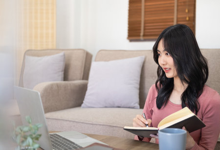 Woman on remote work or online education, using laptop computer, making notes, indoors at home at daytime. Online business, young professional at workplace. Working from home.の写真素材