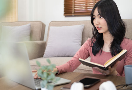 Young woman in casual at wooden table in living room and holding notebook . In front of her is...