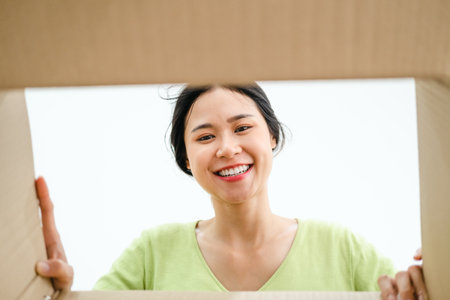 Young pretty woman sitting on couch in living room and opening carton box. Cheerful girl getting parcel at home. Mail delivery to house.の写真素材