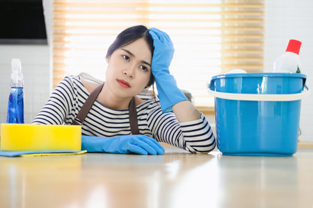 tired woman cleaner in apron and gloves for cleaning sitting with bucket for cleaning on kitchen background, sad girl housewife cleans on house background, cleaning service workerの写真素材