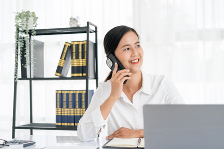 Young asian smiling business woman sit in home office using laptop computer taking by mobile phone. Happy businesswoman sitting at desk behind laptop and talking  on  mobile phone. working from home.の写真素材
