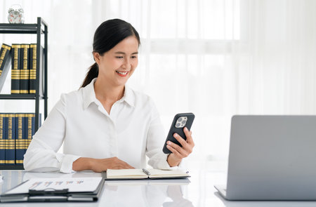 Happy senior woman using mobile phone while working at home with laptop. Smiling mature woman messaging with smartphone. Beautiful stylish elderly lady browsing site on cellphone.の写真素材