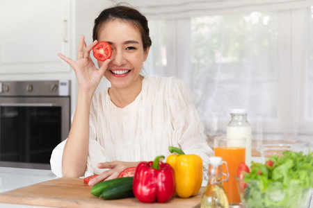 Happy young latin lady having fun and covering eyes with tomato halves, fooling around in kitchen interior, looking and smiling at camera while cooking dinnerの写真素材