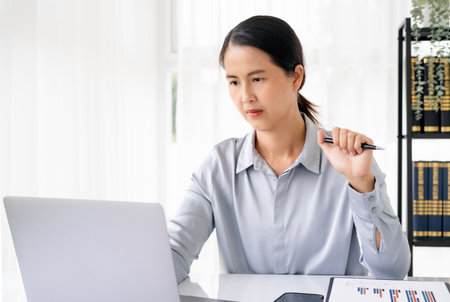 Confident young woman in smart casual wear working on laptop sitting near window in office or cafe. Concentrated at work.の写真素材