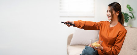 An attractive, positive young Asian woman in comfy clothes is watching TV on a couch in the living room, holding a TV remote control, and changing the TV channel. people and domestic life conceptsの写真素材