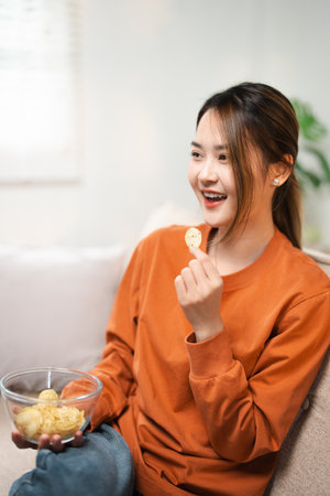 Relaxed asian woman spending free time at home, eating snack and watching TV. Excited smiling lady...
