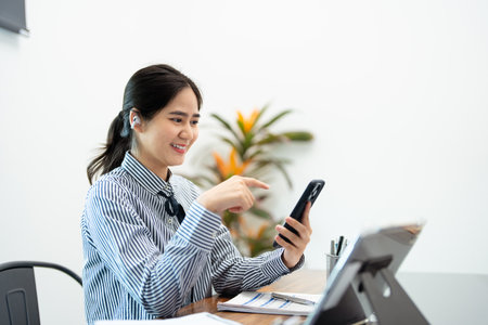 Young busy Asian business woman leader holding cellphone device using mobile phone, looking at smartphone checking financial market app working in modern corporate office .の写真素材