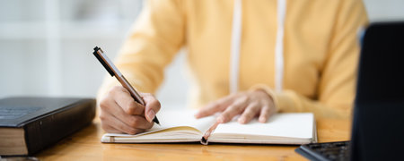 Woman hand with pen writing on notebook with laptop computer on office table. Student studying online class and taking note, e-learning, business event planning, to do list reminderの写真素材
