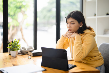 Millennial Asian student prepares for test or exam browses laptop studies in coffee house makes notes in notepad watches online webinar learns foreign language. E learning and online studyingの写真素材