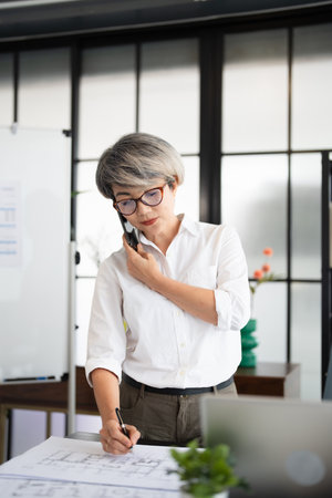 Happy senior woman using mobile phone while working at home with laptop. Cool mature woman  talking with smartphone. Beautiful stylish elderly lady browsing site on cellphone.の写真素材