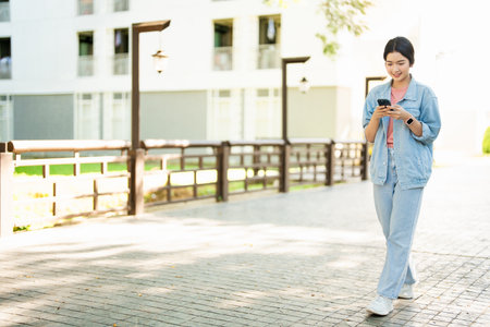 pretty young asian woman walking on walk way holding phone in hands. Use technology concept woman smiling happy using her mobile phone, concept of technology of communication and modern lifestyle.の写真素材