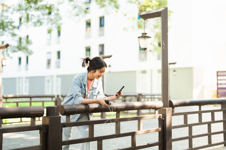 Woman charging her phone with a power bank while standing outdoors on a summer day. Travel device charging concept.の写真素材