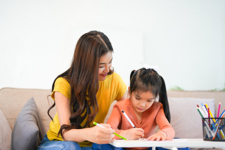 Happy asian girl painting with crayon and colored pencil with her mother in living room at home. Mom teaching daughter how to painting with crayon color on book or doing homework. family concept.の写真素材