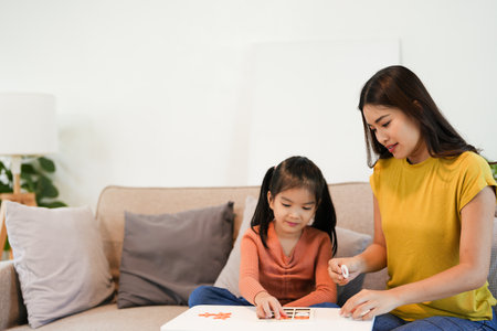 Smiling mother and little daughter playing board game at living room table together, caring mom teaching adorable kid girl, happy family engaged in educational activity at homeの写真素材
