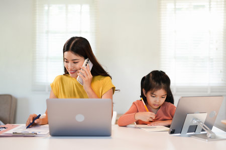 Concept of work from home and home family education. Mom and daughter are sitting at the desk. Business woman works on the Internet in a laptop, a child writes in a notebook.の写真素材