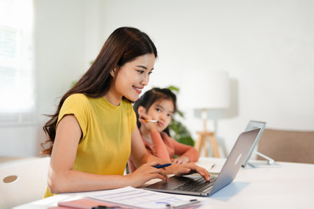 Happy mother working on a computer while her daughter is writing in notebook and learning at home.の写真素材
