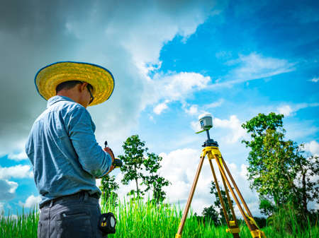 Asian smart engineer or surveyor in black jeans and long sleeve shirt and woven bamboo hat. He is working on controller screen for surveying land in rice field, Thailand. GPS surveying instrument.の写真素材