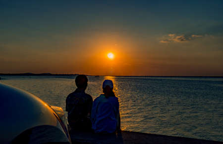 Silhouette of happiness couple sitting and relaxing on the beach in front of the car with orange and...