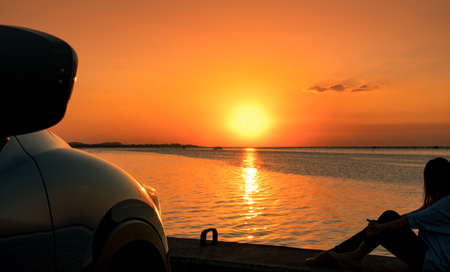 Lonely young woman wear a cap relaxing on the beach alone in front of the car with orange and blue sky at sunset. Summer vacation and travel concept.の写真素材