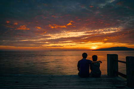 Silhouette of young romantic couple in love is sitting and hugging on wooden pier at the beach in sunrise time with golden sky. Vacation and travel concept. Romantic young couple dating at seaside.の写真素材