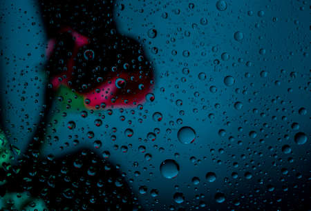Blurred image of a red rose behind a clear glass of a window with a drop of water on blue background with copy space for text. Valentines day concept.の写真素材