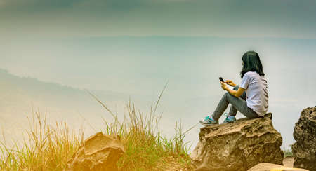 Young Asian woman sit on the rock at the cliff and using smart phone message to her friends while on vacation at the national park. Adventure travel and communication conceptの写真素材