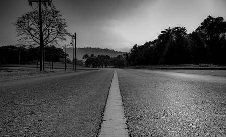 Black and white picture of asphalt road in countryside and the forest beside the road. White strip traffic sign direction paint on asphalt road. The mountain of rainforest as the background.の写真素材