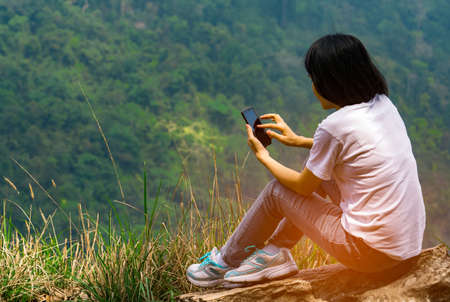 Young Asian woman sit on the rock at the cliff and using smart phone message to her friends while on vacation at the national park. Adventure travel and communication conceptの写真素材