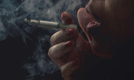 Woman with red nail is smoking cigarette on dark background. Quit smoking concept. Bad habit in...