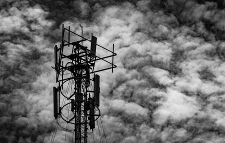 Black and white picture of telecommunication tower against grey sky and white clouds. Antenna on dark sky background. Radio and satellite pole. Communication technology.の写真素材