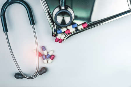 Green stethoscope with pile of antibiotic capsule pills on white table near drug tray. Antimicrobial drug resistance and overuse. Medical equipment for doctor. Clinical Cardiology concept. Health careの写真素材