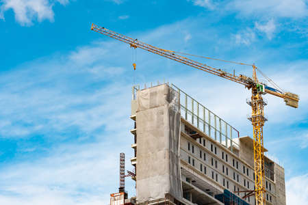 Construction site with crane and building. Real estate industry. Crane use reel lift up equipment in construction site. Building made of steel and concrete. Crane work against blue sky and white cloudの写真素材