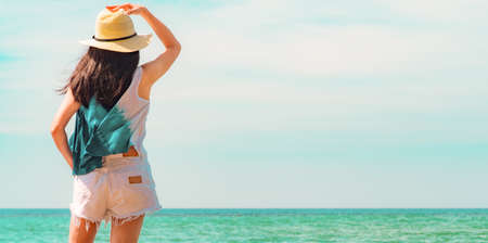 Happy young woman in casual style fashion and straw hat standing at sea beach. Relaxing and enjoy...