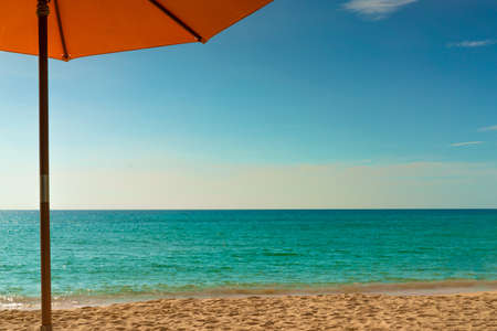 Orange beach umbrella on golden sand beach by the sea with emerald green sea water and blue sky and white clouds. Summer vacation on tropical paradise beach concept. Skyline between sea and sky.の写真素材