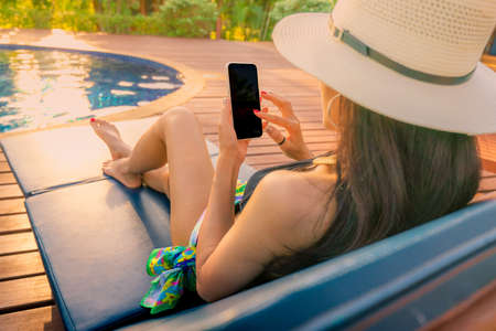 Asian woman with hat and swimsuit sitting in chair at poolside and using smartphone on summer...