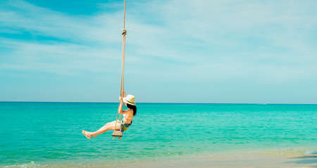 Happy young Asian woman in pink swimsuit and straw hat relax and enjoy holiday at tropical paradise beach. Woman fun on swing. Girl in summer vacation fashion. Green sea water and blue sky and clouds.の写真素材