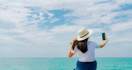 Young Asian woman wear straw hat use smartphone taking selfie at wooden pier. Summer vacation at tropical paradise beach. Happy girl travel on holiday. Woman enjoy and relax life. Summer vibes.の写真素材
