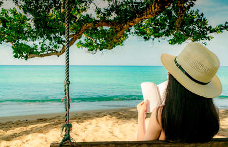 Women sit and reading a book on swings under the tree by the sea. Back view of sexy Asian woman with straw hat relax and enjoy holiday at tropical paradise sand beach. Summer vacation. Summer vibes.の写真素材