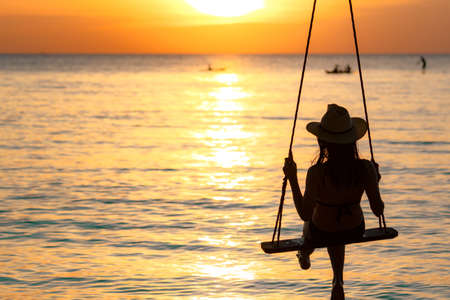 Silhouette woman wear bikini and straw hat swing the swings at the beach on summer vacation at sunset. Girl in swimwear sit on swings and watch beautiful sunset. Summer vibes. Woman travel alone.の写真素材
