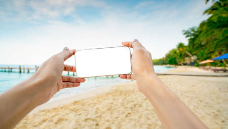 Woman hands holding smartphone with empty screen display on sand beach of tropical island. Woman taking a photo of paradise beach on summer vacation. Adult Asian woman enjoying and relaxing on beach.の写真素材