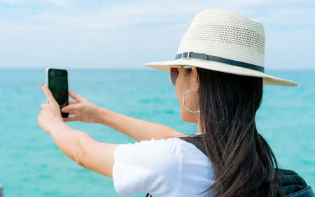 Young Asian backpacker woman wear hat use smartphone taking selfie at pier. Summer vacation at tropical paradise beach. Happy hipster girl travel on holiday. Woman enjoy and relax life. Summer vibes.の写真素材