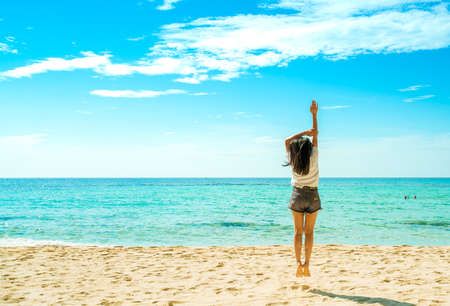 Happy young woman in white shirts and shorts jumping at sand beach. Relaxing and enjoying holiday at tropical paradise beach with blue sky and clouds. Girl in summer vacation. Summer vibes. Happy day.の写真素材